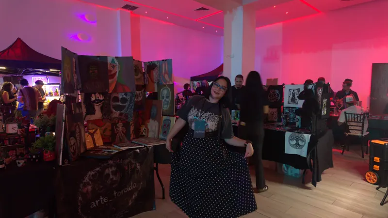 Sara Villa con playera de banda y falda de lunares frente a su stand, el segundo día, con el salón en luz rosa