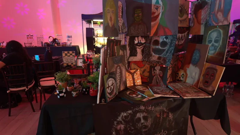 Pared de pinturas del stand de Arte Torcido bajo luz rosa: payasos, máscaras y rostros, con la lona de arte_torcido al frente